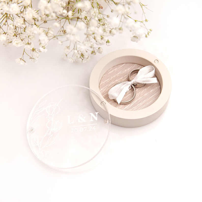 May include: A round, clear acrylic ring box with a floral design and the inscription "L&N 2023.07.24". The box is open, revealing a white ring pillow with two gold wedding bands tied with a white ribbon.