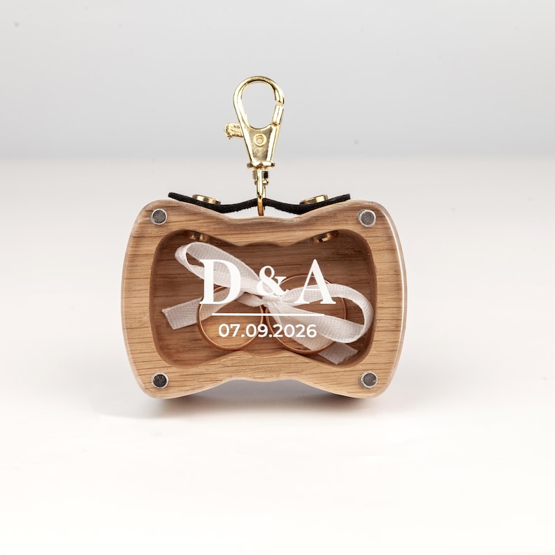 May include: A light wood ring box with a gold-tone metal lobster clasp at the top. The front features white printed text reading D & A and 07.09.2026. Inside, two gold rings are tied together with a white ribbon, secured by four small magnets.