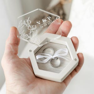 Puede incluir: Una caja hexagonal blanca para anillos con tapa transparente, grabada con diseños florales, iniciales y una fecha. En el interior, dos anillos plateados están atados con una cinta blanca. La caja se sostiene abierta, mostrando los anillos.