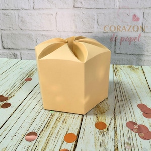 Puede incluir: Una caja de regalo hexagonal beige con un cierre único en forma de pétalo. La caja es de papel y está sobre una superficie de madera salpicada de confeti color cobre. El texto "CORAZON de papel" es visible en el fondo.