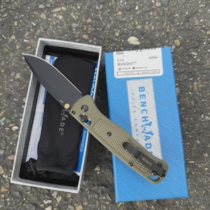 May include: A Benchmade 535 Bugout knife with a black blade and olive green handle, displayed open next to its box. The box is blue with white text, and the knife is in a white box with a black pouch.