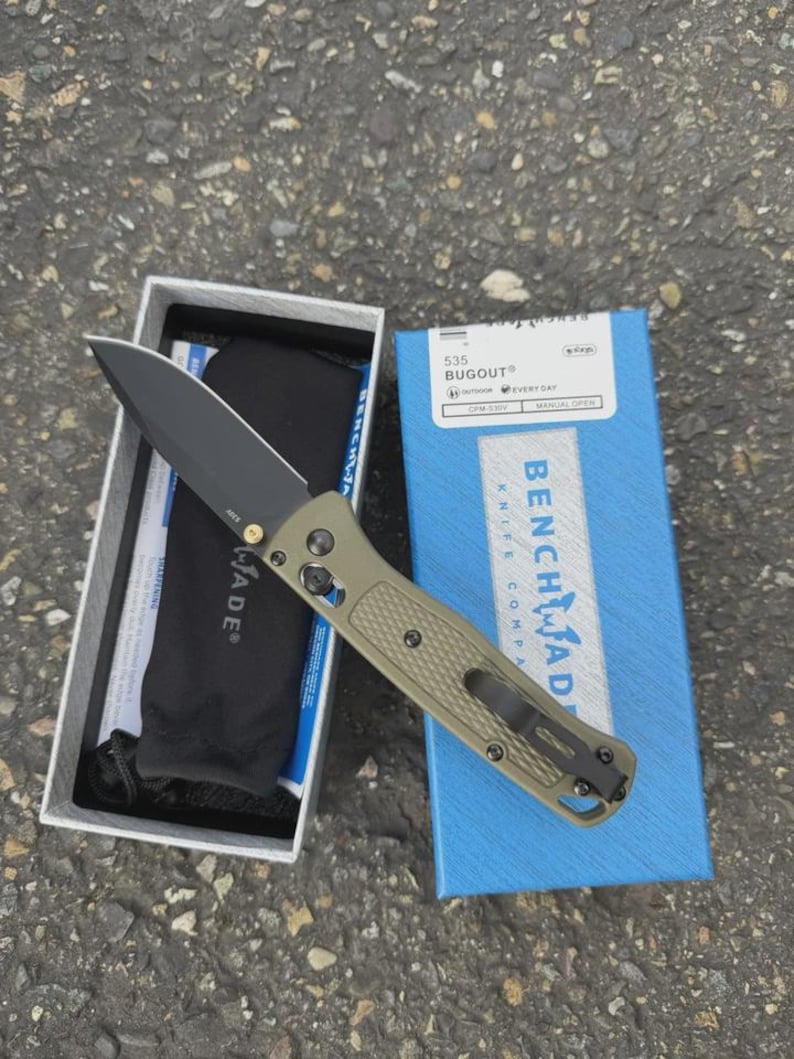 May include: A Benchmade 535 Bugout knife with a black blade and olive green handle, displayed open next to its box. The box is blue with white text, and the knife is in a white box with a black pouch.