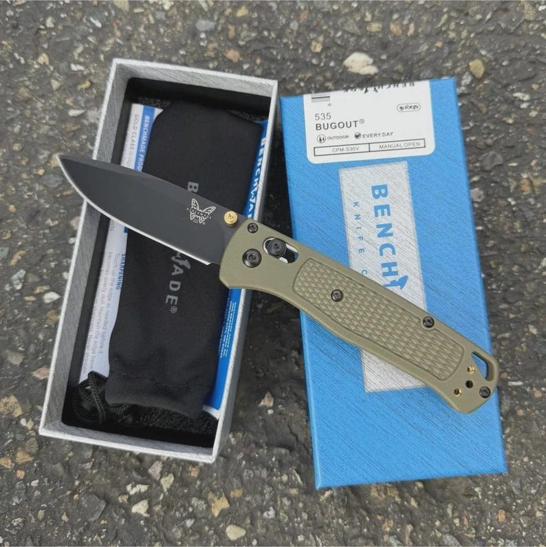 May include: A Benchmade Bugout 535 knife with a black blade and olive green handle, displayed in its original box. The box is blue with white text, and the knife is partially out of its packaging.