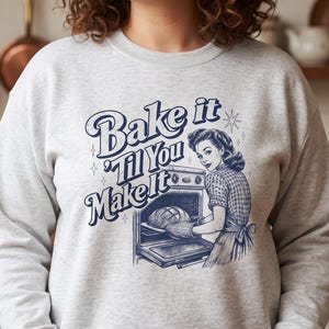 May include: A light gray crewneck sweatshirt featuring a dark blue graphic print. The design displays a person wearing an apron, placing a loaf of bread into an oven, accompanied by the text "Bake it 'Til You Make it" in a retro font style.