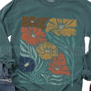 May include: A teal long-sleeved shirt featuring a floral print in orange, yellow, and blue. The shirt is displayed with blue jeans and brown ankle boots. The text "Stella Vita" is visible.