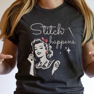 May include: Dark grey t-shirt featuring the phrase "Stitch happens" and a retro illustration of a woman with a needle. The woman has a red bow and earrings. The t-shirt is a crew neck.