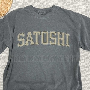 May include: A dark gray t-shirt with the word "SATOSHI" printed in a light beige color on the front. The shirt is made of a soft, comfortable fabric.