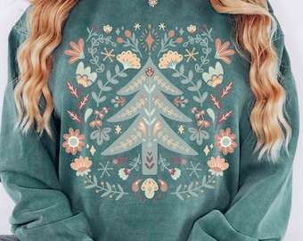 Może przedstawiać: Turkusowa bluza z kolorowym wzorem kwiatowym przedstawiającym stylizowany choinkę. Tekst "Stella Vita" jest widoczny na dole obrazu. Kolor bluzy opisano jako "Blue Spruce".