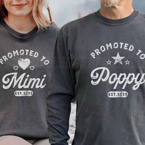 May include: Two long-sleeved shirts with a distressed, vintage look. The shirt on the left says "PROMOTED TO Mimi EST. 2025" with two hearts. The shirt on the right says "PROMOTED TO Poppy EST. 2025" with a star.
