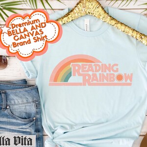 May include: Light blue t-shirt with the text "READING RAINBOW" in a retro design featuring a rainbow and sun. The shirt is a Bella + Canvas brand. The shirt is paired with denim shorts and brown sandals.