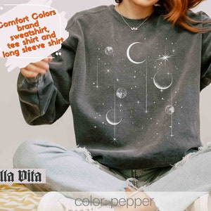 Op de afbeelding: Een grijs sweatshirt met een wit maan- en sterrenontwerp. De tekst "Comfort Colors brand sweatshirt, tee shirt and long sleeve shirt" is gedrukt in de linkerbovenhoek van het sweatshirt. De tekst "Stella Vita" is gedrukt in de linkerbenedenhoek van het sweatshirt. De tekst "color: pepper" is gedrukt in de rechterbenedenhoek van het sweatshirt.