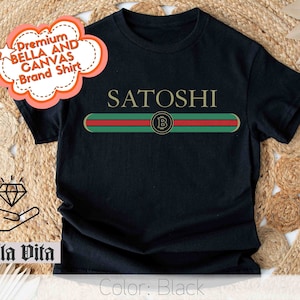 May include: Black t-shirt with a green, red, and white stripe across the chest. The stripe has a gold Bitcoin symbol in the center. The text "SATOSHI" is above the stripe.