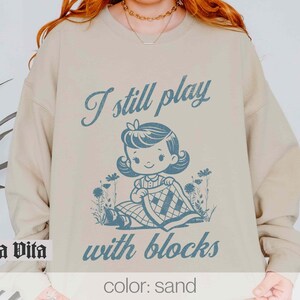 K&ouml;nnte beinhalten: Ein sandfarbenes Sweatshirt mit dem Text "I still play with blocks" in Blau. Eine Cartoon-Illustration eines M&auml;dchens, das mit Bl&ouml;cken spielt, ist mittig auf dem Sweatshirt platziert.