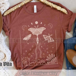 May include: A heather clay-colored t-shirt featuring a detailed beige graphic of a moth with moon phases, butterflies, and floral elements. The text "Stella Vita" is visible on the shirt. The shirt is folded, with a hat and jeans in the background.