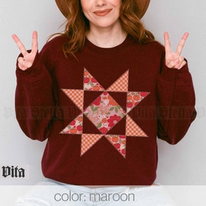 Puede incluir: Una sudadera color granate con un gran diseño de estrella de patchwork colorido. La estrella está hecha de varios tonos de rosa, naranja y rojo. El texto "color: maroon" está impreso en la parte inferior de la imagen.