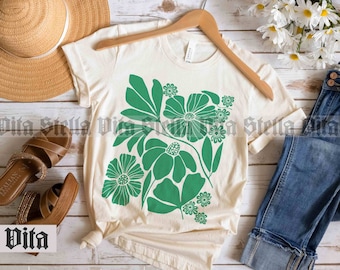 May include: A natural colored t-shirt with a green floral design. The shirt is laid flat on a wooden surface with a straw hat, sandals, and blue jeans.