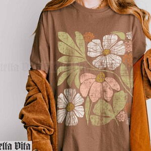 May include: A brown t-shirt with a vintage floral print featuring a large white flower, a pink flower, and green leaves. The print has a distressed look.