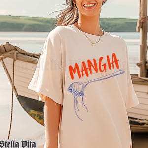 May include: A white t-shirt with the word "MANGIA" printed in orange and a blue and white illustration of a fork with spaghetti on it.