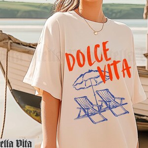 May include: A white t-shirt with the text "Dolce Vita" in red and blue lettering. The shirt features a graphic of two beach chairs and an umbrella.