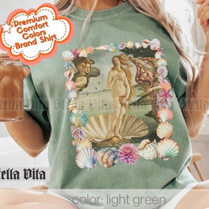 Puede incluir: Una camiseta verde claro con un estampado gr&aacute;fico de un borde de conchas que rodea una pintura de una mujer de pie sobre una concha. El texto "color: light green" est&aacute; impreso en la parte inferior de la imagen.