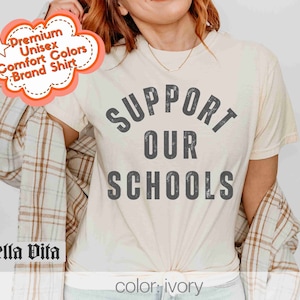 May include: A white t-shirt with the text "SUPPORT OUR SCHOOLS" in a distressed gray font. The shirt is being modeled by a person wearing a plaid shirt.