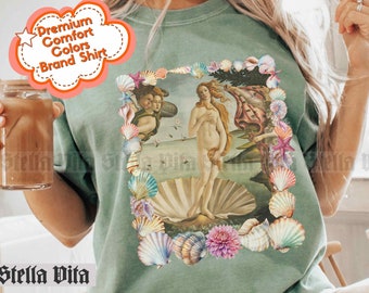Puede incluir: Una camiseta verde claro con un estampado gráfico de un borde de conchas que rodea una pintura de una mujer de pie sobre una concha. El texto "color: light green" está impreso en la parte inferior de la imagen.