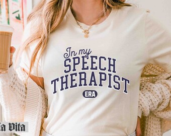 Può includere: Una maglietta bianca con il testo "In my Speech Therapist ERA" stampato in blu navy. Il testo è in uno stile di font universitario.
