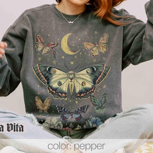 May include: A charcoal grey sweatshirt with a graphic print of a large moth with wings spread wide, surrounded by smaller butterflies and a crescent moon. The text "Stella Vita" is printed on the bottom left corner of the sweatshirt.