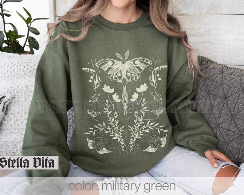 May include: A green sweatshirt with a white graphic of a moth and flowers. The text "Stella Vita" is printed on the bottom left corner of the sweatshirt. The text "color: military green" is printed on the bottom right corner of the sweatshirt.