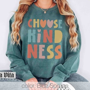 K&ouml;nnte beinhalten: Ein blaugr&uuml;ner Pullover mit dem Text "Choose Kindness" in bunten Buchstaben mit Herzen. Die Buchstaben sind in einer Schriftart im Retro-Stil.