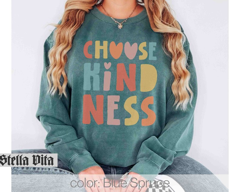 K&ouml;nnte beinhalten: Ein blaugr&uuml;ner Pullover mit dem Text "Choose Kindness" in bunten Buchstaben mit Herzen. Die Buchstaben sind in einer Schriftart im Retro-Stil.