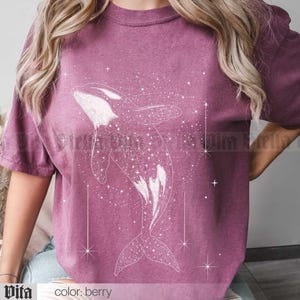 May include: A berry colored t-shirt with a white outline of an orca whale swimming through a field of stars. The text "Stella Vita" and "color: berry" are at the bottom of the image.