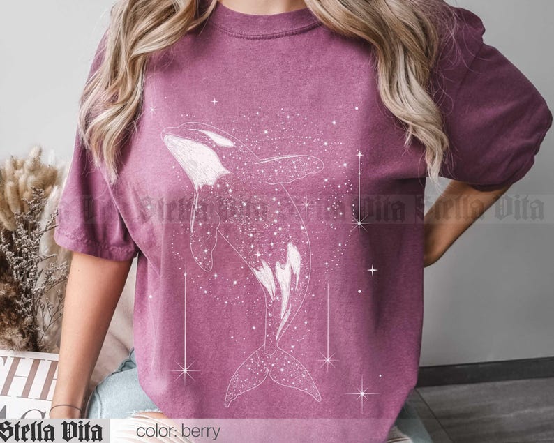 May include: A berry colored t-shirt with a white outline of an orca whale swimming through a field of stars. The text "Stella Vita" and "color: berry" are at the bottom of the image.