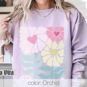 May include: A lavender sweatshirt with a floral design featuring pink, yellow, and green flowers. The flowers have hearts in the centre. The text "color: Orchid" is printed on the bottom of the sweatshirt.
