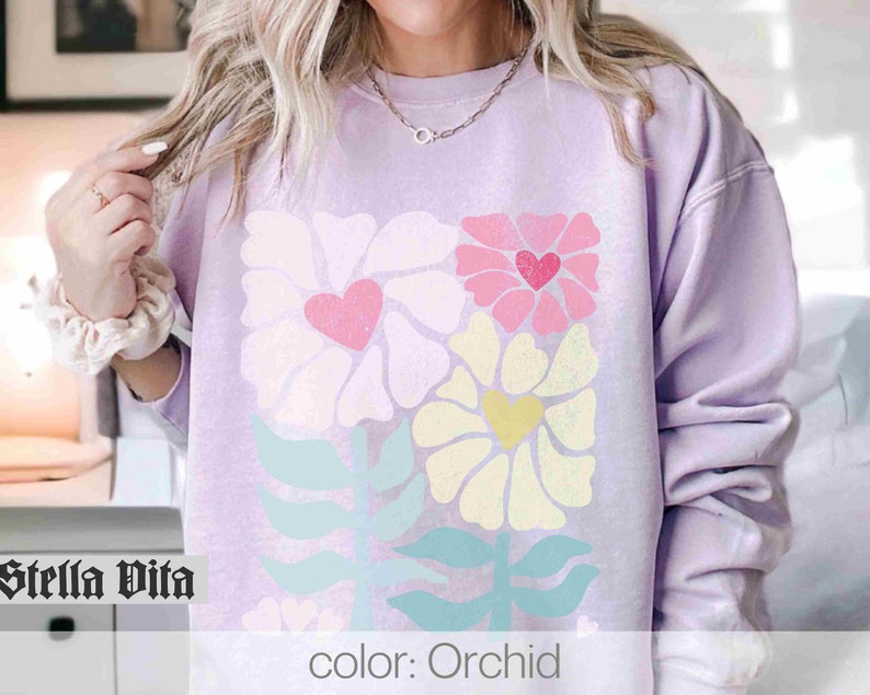 May include: A lavender sweatshirt with a floral design featuring pink, yellow, and green flowers. The flowers have hearts in the centre. The text "color: Orchid" is printed on the bottom of the sweatshirt.