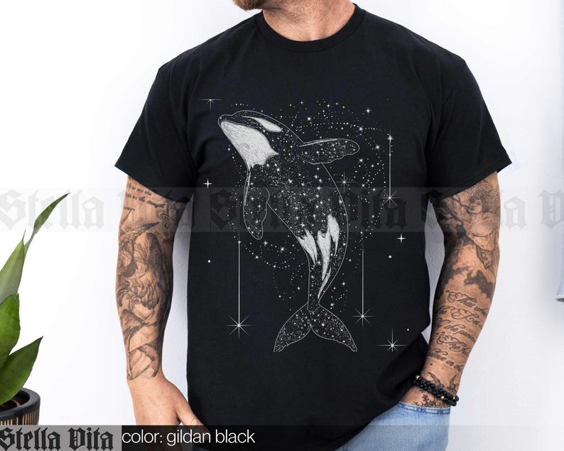 May include: Black t-shirt featuring a white graphic of an orca whale swimming in a field of stars. The text "Stella Vita" is printed on the bottom of the shirt.