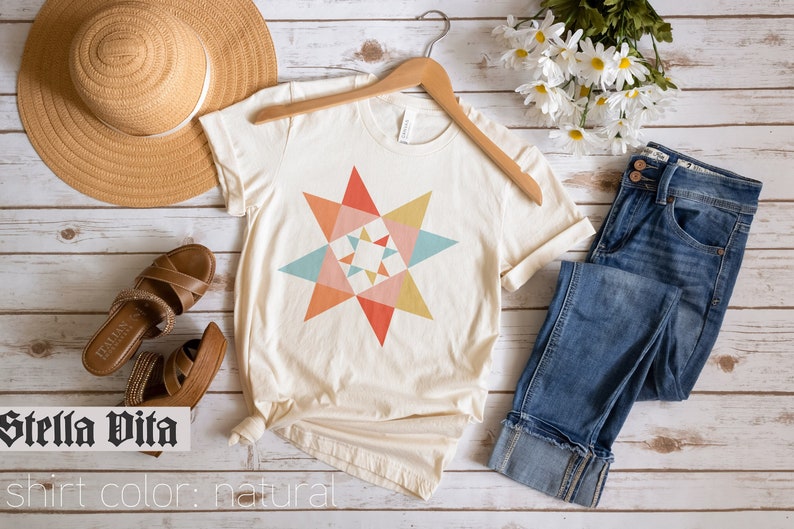 May include: A natural colored t-shirt with a colorful geometric star design. The shirt is on a hanger and there are jeans, sandals, a hat, and flowers in the background. "Stella Vita" and "shirt color: natural" are written below the shirt.
