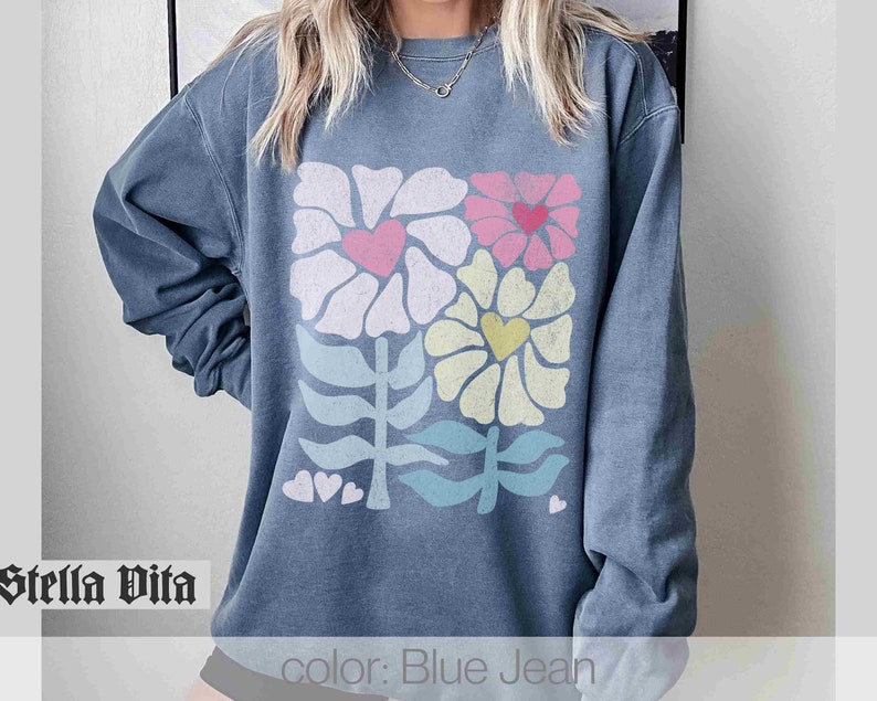 May include: A blue jean sweatshirt with a colourful floral design featuring hearts and leaves. The text "Stella Vita" is visible in the bottom left corner of the image. The text "color: Blue Jean" is visible in the bottom right corner of the image.