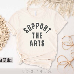 May include: A natural colored t-shirt with the text "SUPPORT THE ARTS" in a distressed black font.