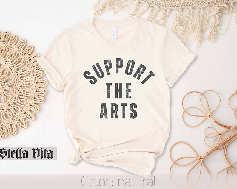 May include: A natural colored t-shirt with the text "SUPPORT THE ARTS" in a distressed black font.