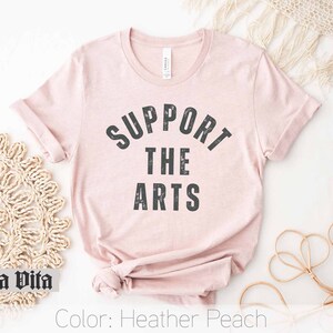May include: A heather peach t-shirt with the text "Support the Arts" in black lettering.