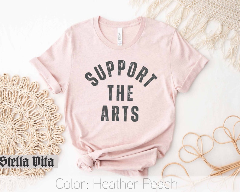 May include: A heather peach t-shirt with the text "Support the Arts" in black lettering.
