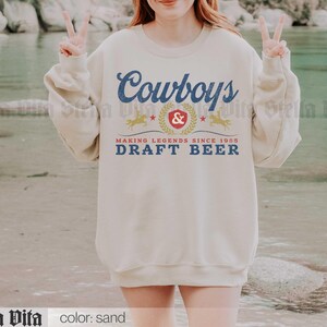 May include: Sand-colored sweatshirt with the text "Cowboys & Draft Beer" in blue and gold. The design includes a crest, stars, and horse silhouettes, with the words "Making Legends Since 1985".
