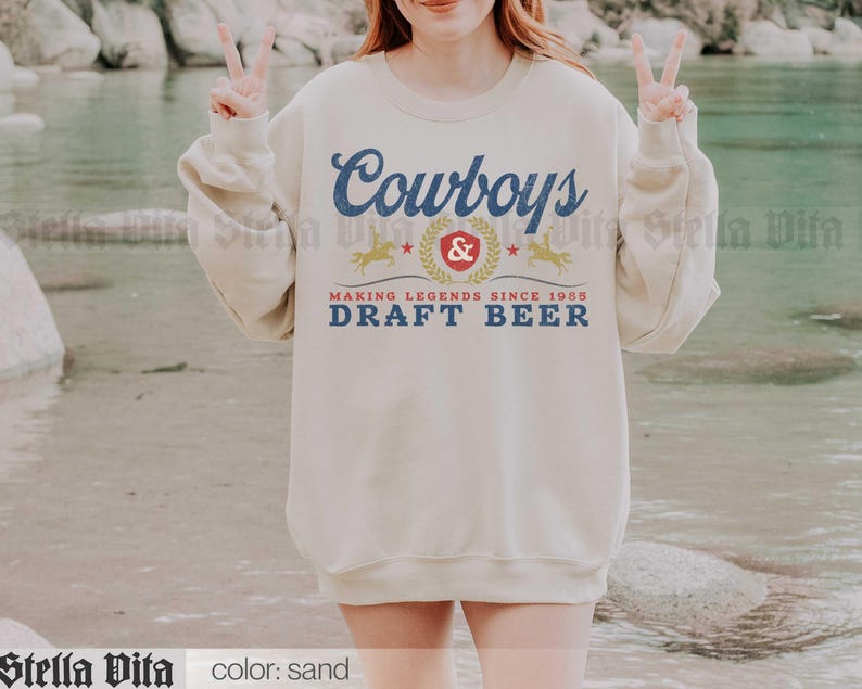 May include: Sand-colored sweatshirt with the text "Cowboys & Draft Beer" in blue and gold. The design includes a crest, stars, and horse silhouettes, with the words "Making Legends Since 1985".