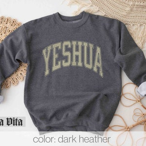 May include: Dark heather grey sweatshirt with the word "YESHUA" printed in a vintage-style, arched font.