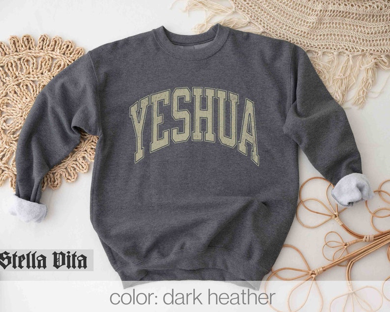 May include: Dark heather grey sweatshirt with the word "YESHUA" printed in a vintage-style, arched font.