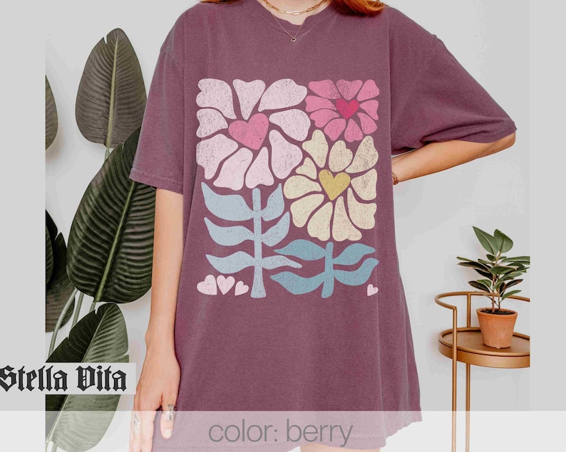 May include: A purple t-shirt featuring a graphic print of stylised flowers and hearts in pink, yellow, blue and green. The text "Stella Vita" is visible in the bottom left corner of the image. The text "color: berry" is visible in the bottom right corner of the image.