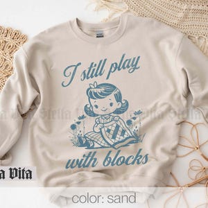 K&ouml;nnte beinhalten: Ein beiger Pullover mit dem Text "I still play with blocks" in blauer Schrift. Unter dem Text befindet sich eine Comic-Illustration eines M&auml;dchens, das mit Bl&ouml;cken spielt. Der Pullover ist in der Farbe Sand erh&auml;ltlich.
