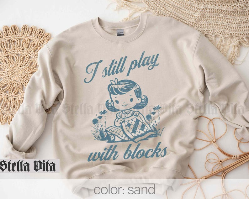 K&ouml;nnte beinhalten: Ein beiger Pullover mit dem Text "I still play with blocks" in blauer Schrift. Unter dem Text befindet sich eine Comic-Illustration eines M&auml;dchens, das mit Bl&ouml;cken spielt. Der Pullover ist in der Farbe Sand erh&auml;ltlich.