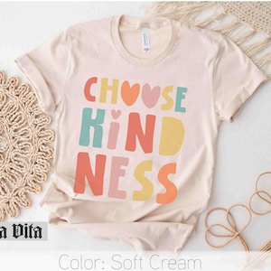 K&ouml;nnte beinhalten: Ein T-Shirt in zartem Creme mit dem Text "Choose Kindness" in bunten Blockbuchstaben mit Herzen.